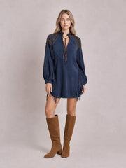 Model stands facing forward in a navy mini dress with lantern sleeves, shoulder detailing, a V-neck tie, and long fringe along the sides and hem.