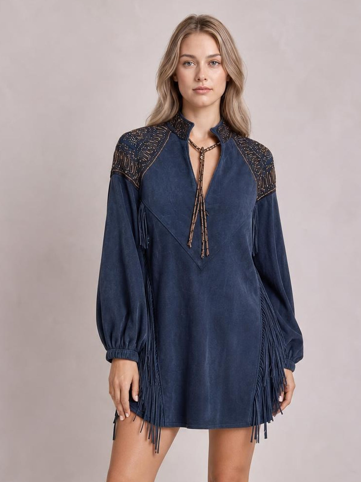 Model standing in a navy mini dress with voluminous lantern sleeves, a high collar, patterned shoulder panels, lace-up neckline, and long fringe trim.