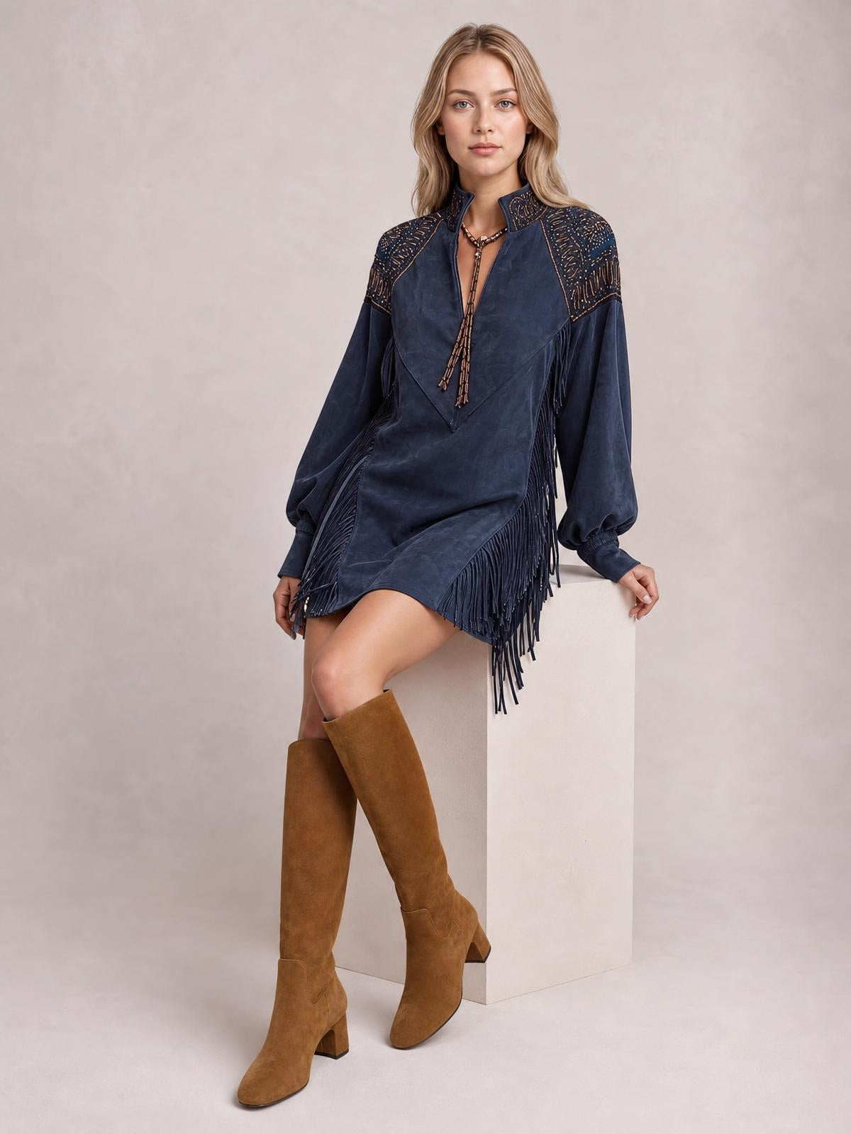 Model poses seated in a navy fringed lantern sleeve mini dress with a relaxed silhouette, embroidered shoulder panels, and long fringe along the sides and hem.