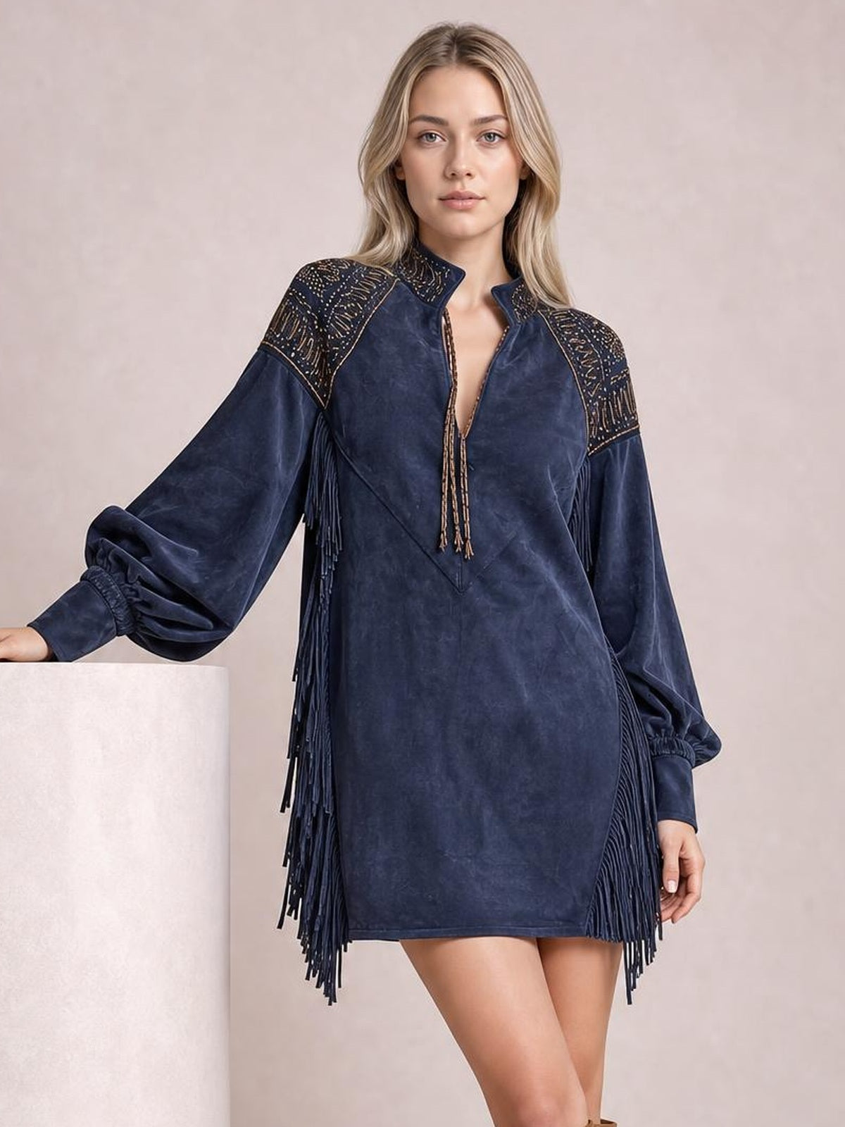 Model poses with one hand on a pedestal in a navy fringed mini dress featuring lantern sleeves, embellished shoulders, and a front zipper.