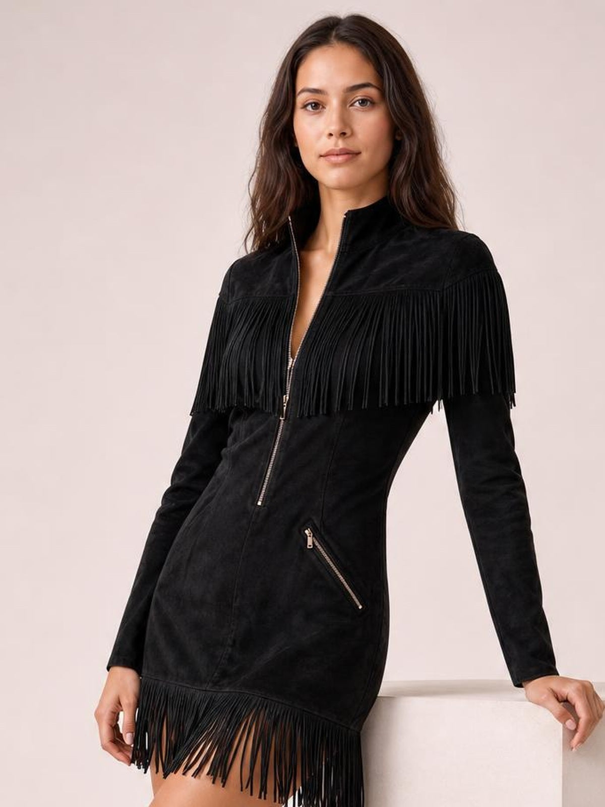 A model leans against a surface in a black fringed zip-front mini dress with long sleeves, a stand collar, diagonal zip pocket, and fringe trim.