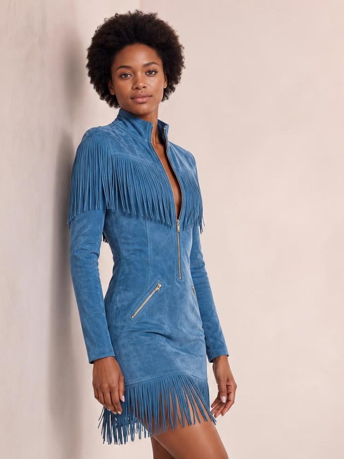 Model poses in a blue fringed zip-front mini dress with long sleeves, a fitted silhouette, tassel-trimmed shoulders and hem, and diagonal zip pockets.