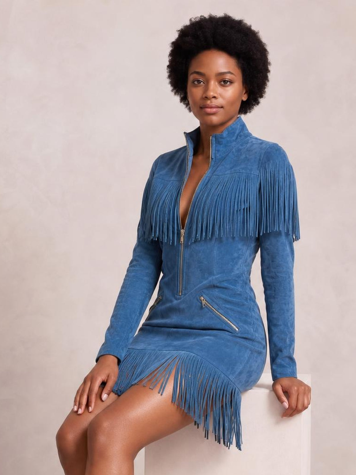 A seated model wears a blue zip-front mini dress with long sleeves, a stand collar, diagonal zip pockets, and dramatic fringe trim across the chest and hem.