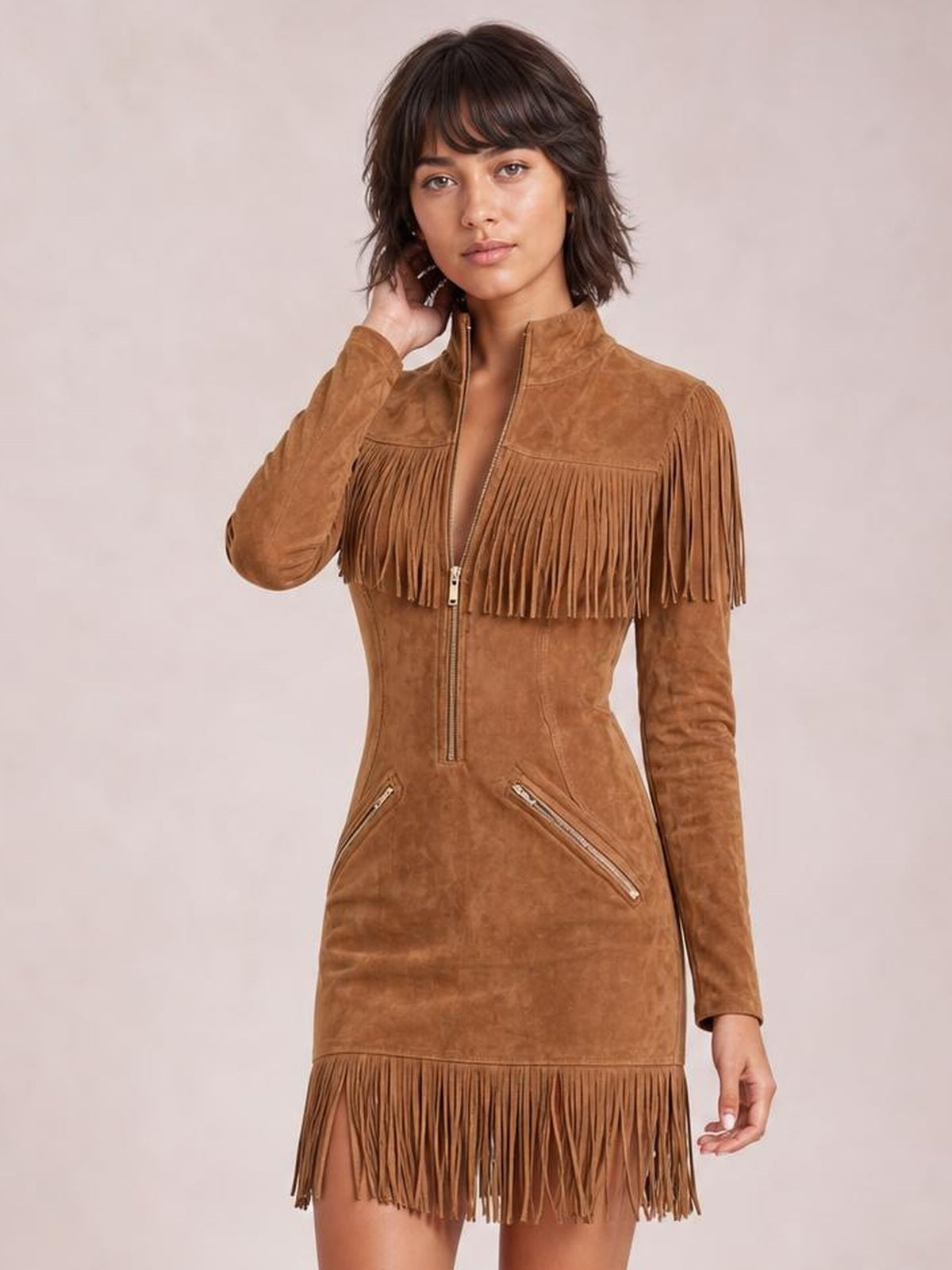Model poses with one hand in her hair wearing a fitted tan mini dress with long sleeves, zip front, angled zip pockets, and fringe trim.