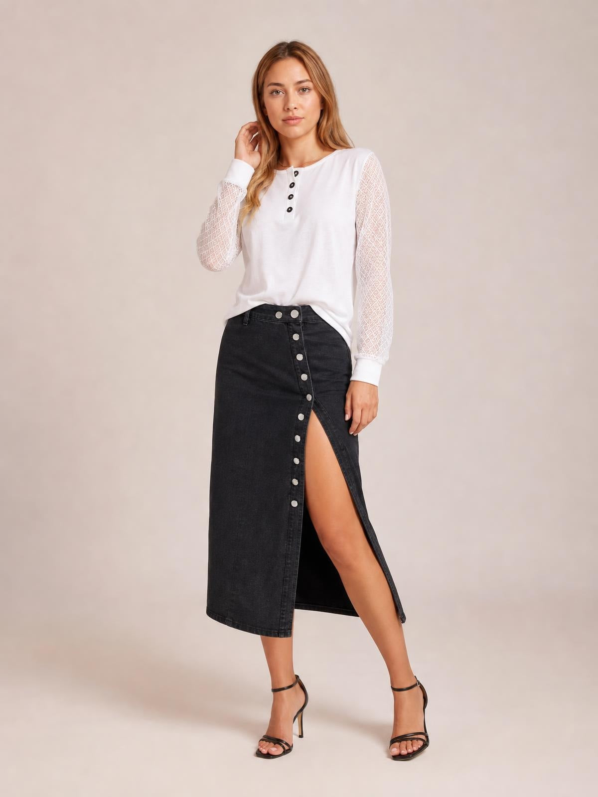 Model poses in a black denim midi skirt with a straight silhouette, front button closure, and a high front slit, paired with a white top.