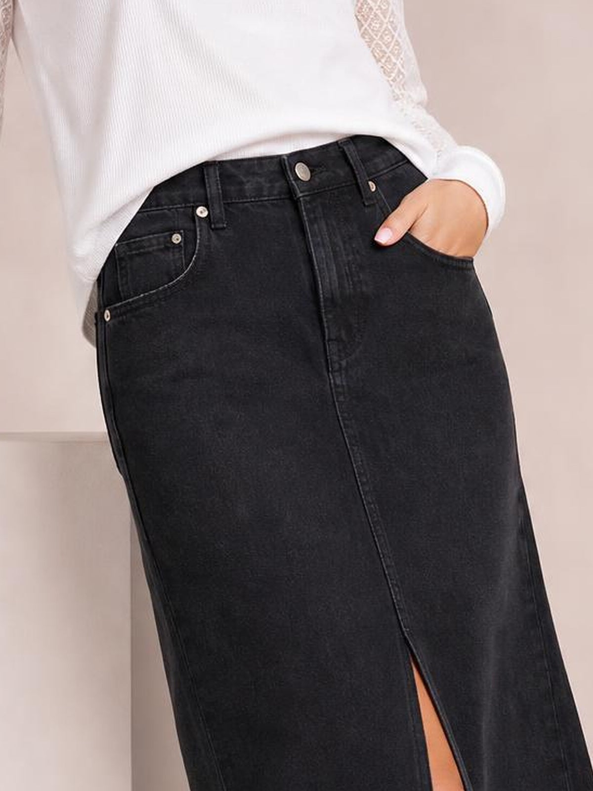 A model wears a black front-button midi denim skirt with belt loops, pockets, a straight silhouette, and a front slit, styled with a white top.