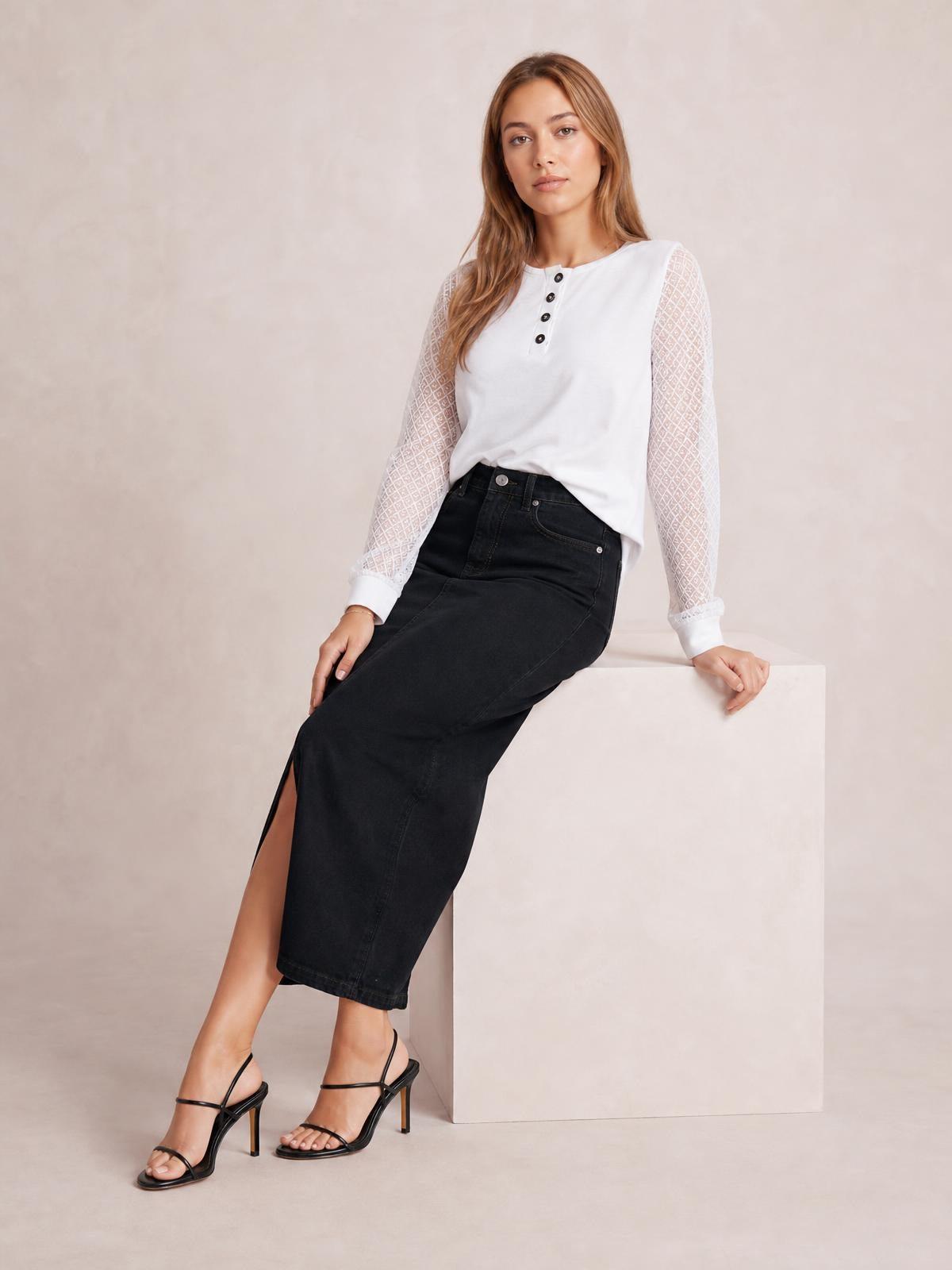 Model seated in a black front-button slit midi denim skirt with a high waist, fitted silhouette, and visible front seam construction.