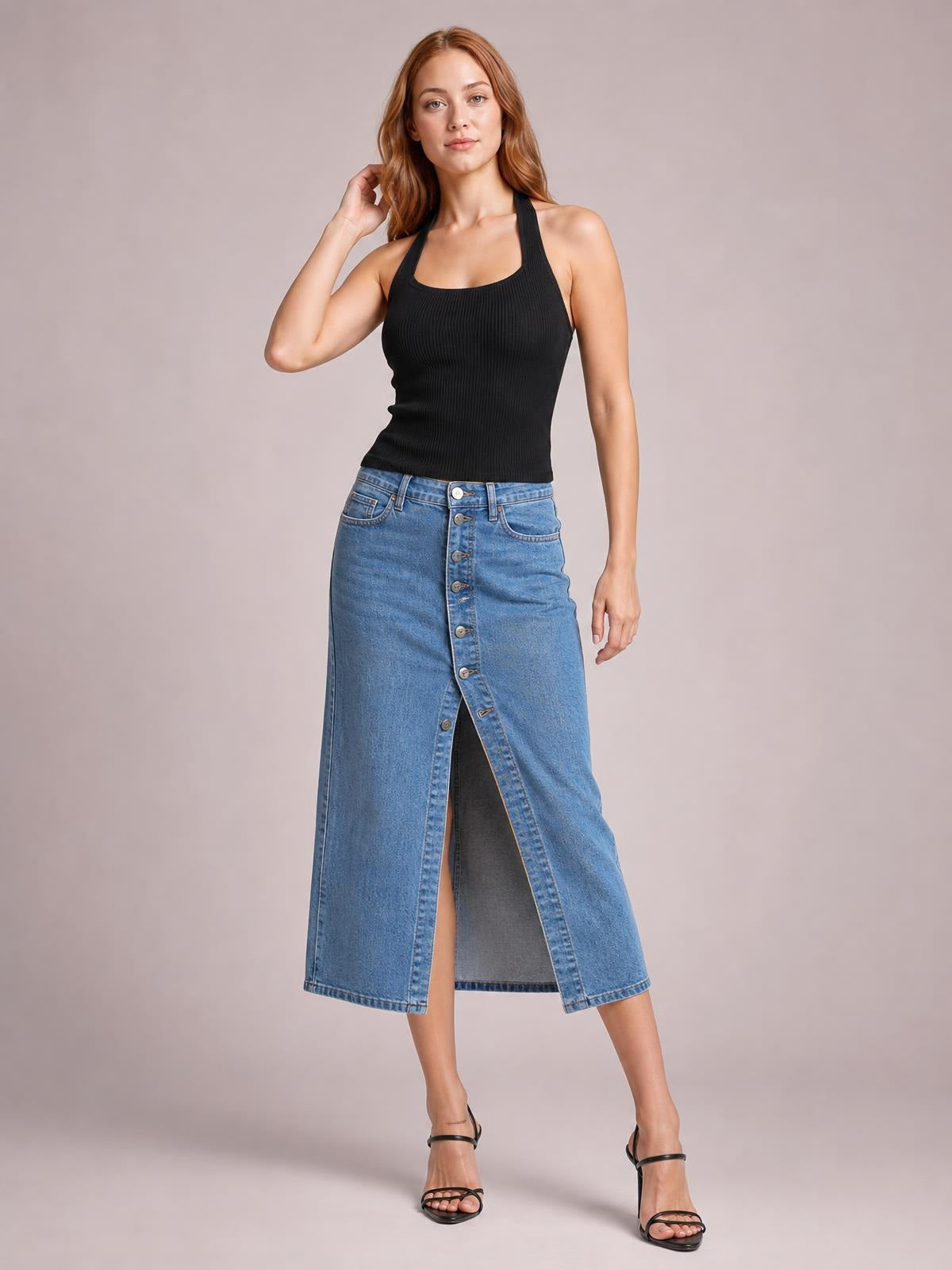 Model poses with one hand in her hair wearing a blue denim midi skirt with a straight silhouette, front button closure, and dramatic center slit.