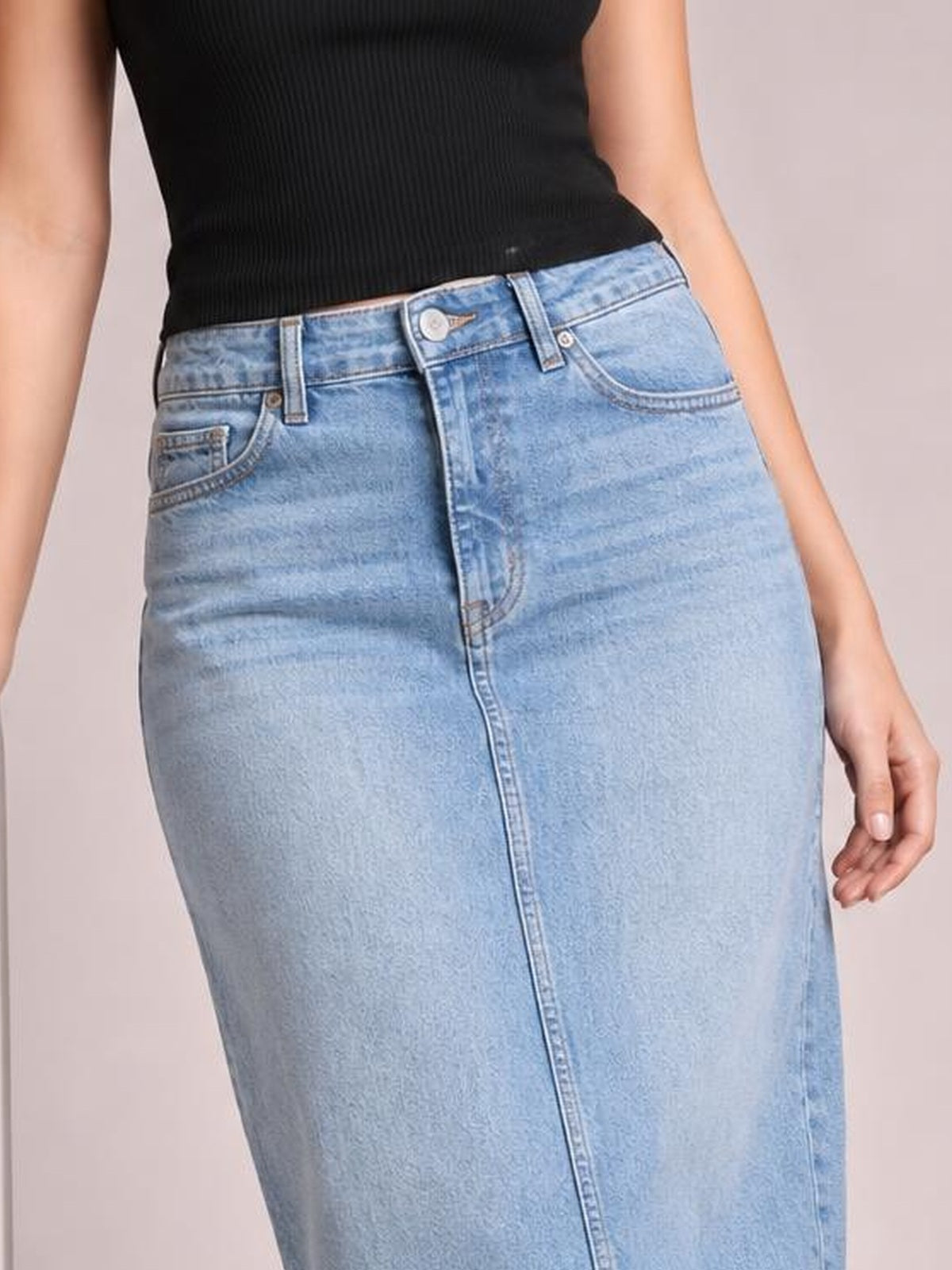 A model wears a light blue denim midi skirt with a fitted straight silhouette, belt loops, front pockets, and a buttoned waistband, shown from the waist down.