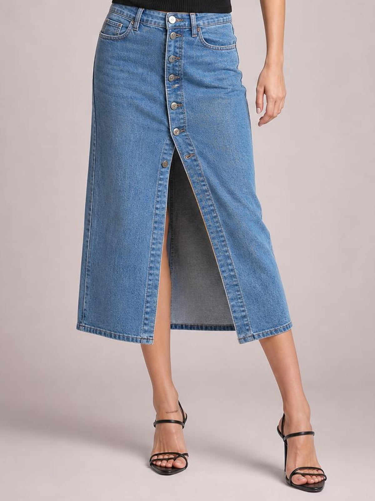 Model stands in a blue denim midi skirt with a front button closure, high waist, straight silhouette, and front slit, paired with black heeled sandals.