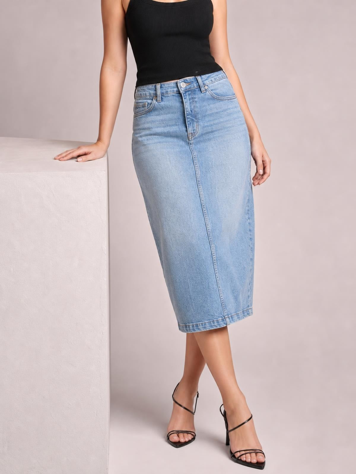 A model wears a light blue front-button denim midi skirt with a fitted straight silhouette, belt loops, pockets, and a crossed-leg pose.