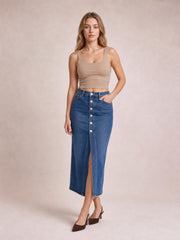 Model wearing a blue denim midi skirt with a front button closure, center slit, fitted silhouette, and raw hem, posing with one hand in a pocket.