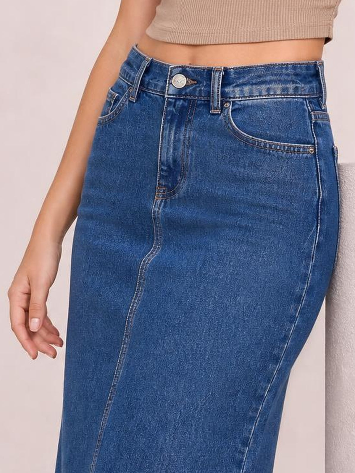 A model wears a blue denim midi skirt with a fitted straight silhouette, front button closure, belt loops, contrast stitching, and curved front pockets.