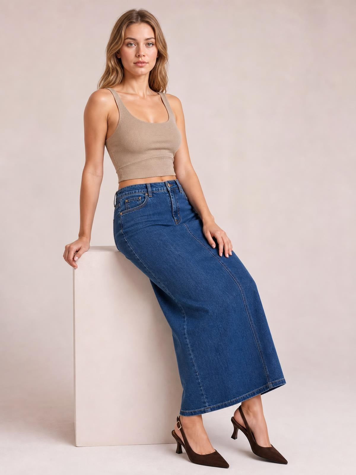 A model leans against a block wearing a blue denim midi skirt with a high waist, front buttons, stitched panels, and a slim straight silhouette.