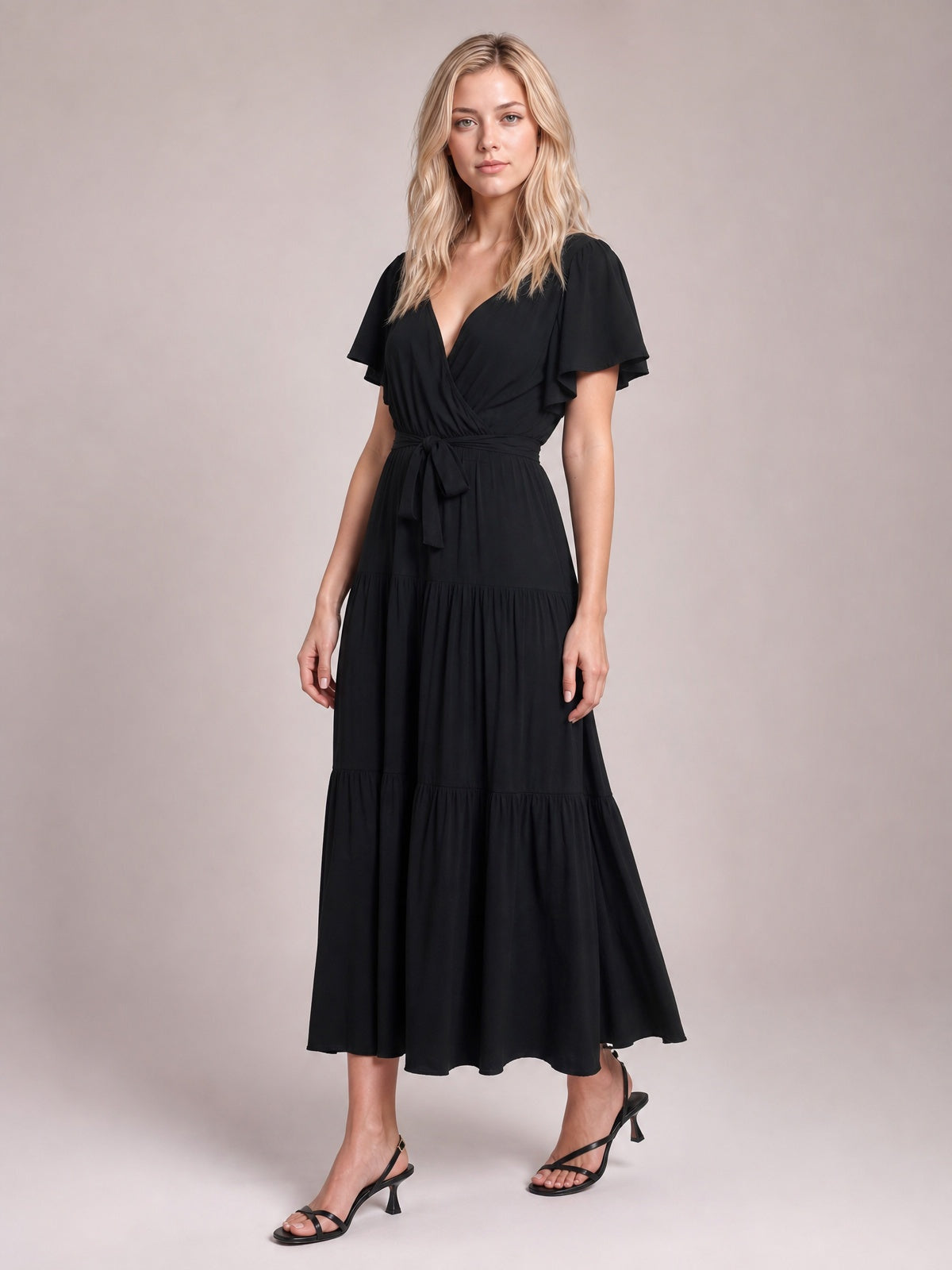 A model stands in a black front-tie midi dress with a V-neck, flutter short sleeves, defined waist, and flowing tiered skirt.