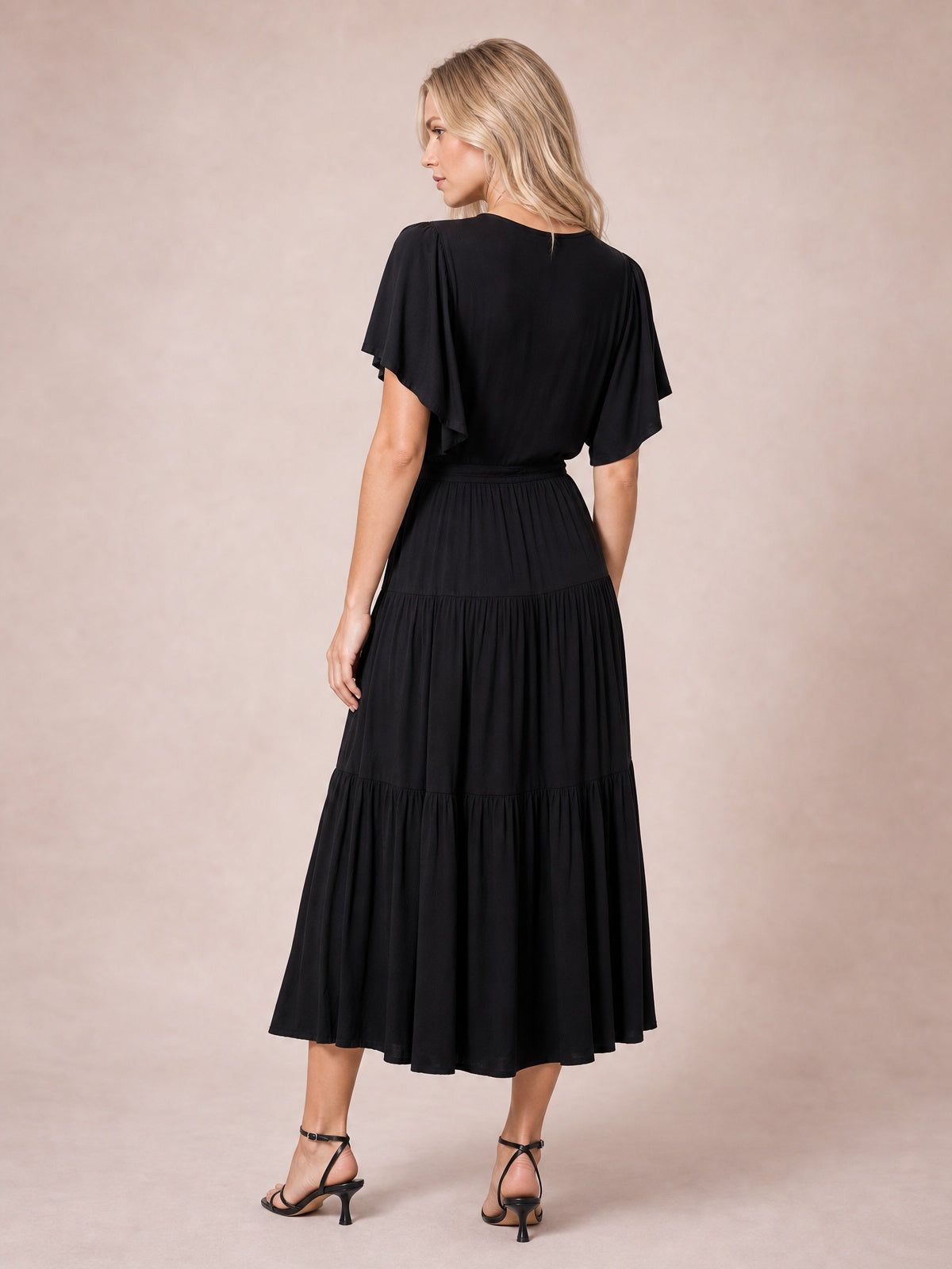 A model poses turned away in a black short sleeve tiered midi dress with flutter sleeves, a gathered waist, and a flowing tiered skirt.