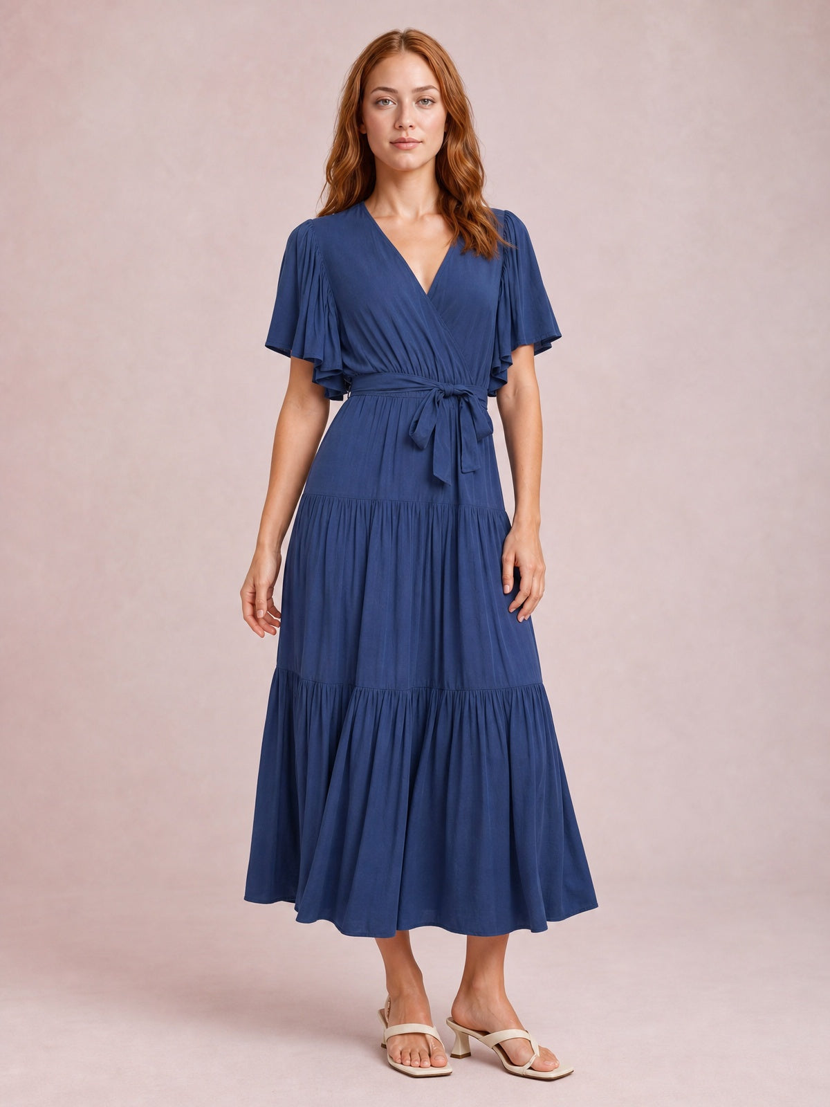 A model stands in a navy blue midi dress with a V-neck, flutter short sleeves, front waist tie, and flowing tiered skirt.