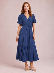 A model stands in a navy blue midi dress with a V-neck, flutter short sleeves, front waist tie, and flowing tiered skirt.
