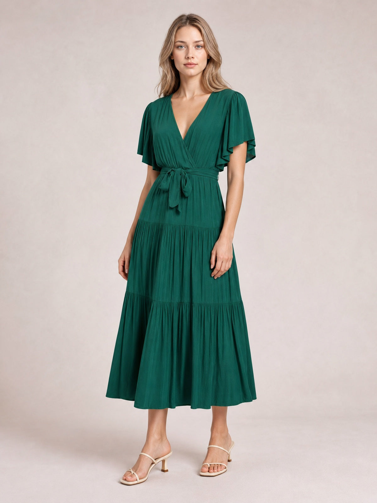 Model wearing a deep green short-sleeve tiered midi dress with a V-neckline, front waist tie, flutter sleeves, and flowing gathered skirt.