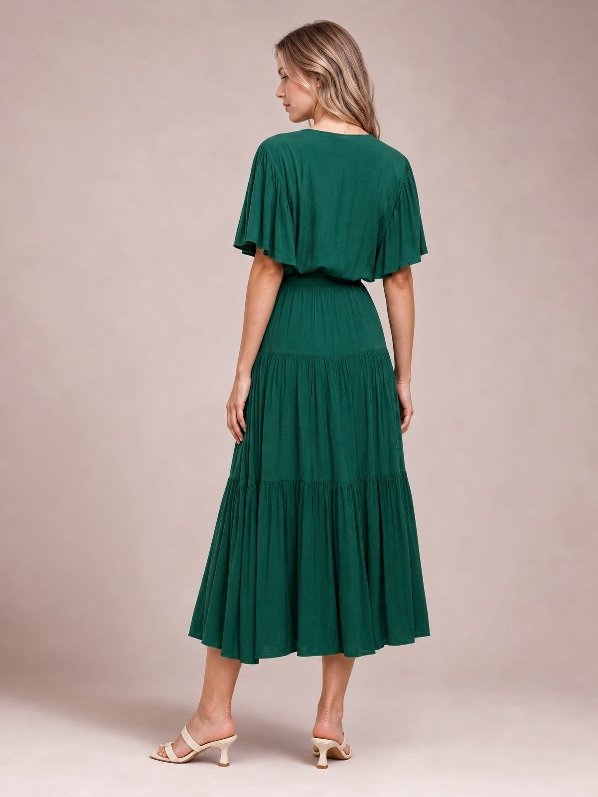 A model looks over her shoulder in a deep green tiered midi dress with flutter short sleeves, a defined waist, and a flowing gathered skirt.