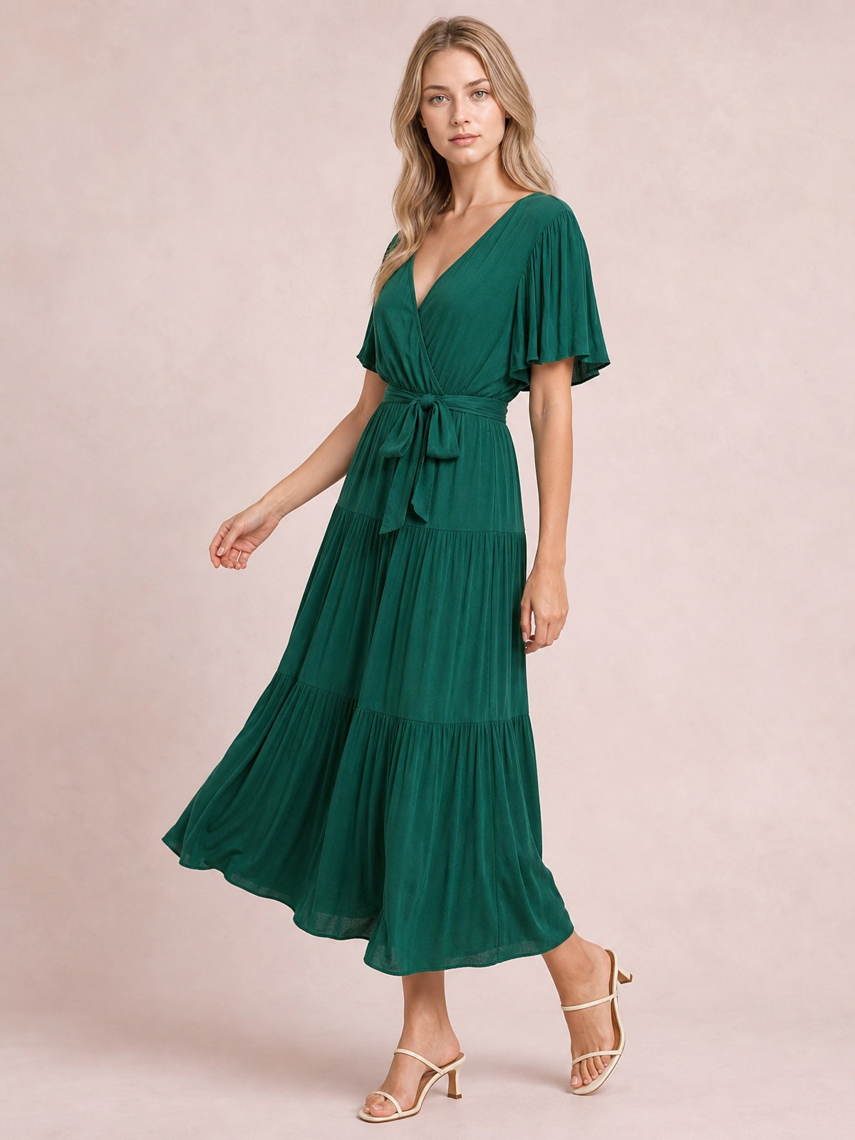 Model wears a deep green front-tie midi dress with a V-neckline, flutter short sleeves, a defined waist, and a flowing tiered skirt.