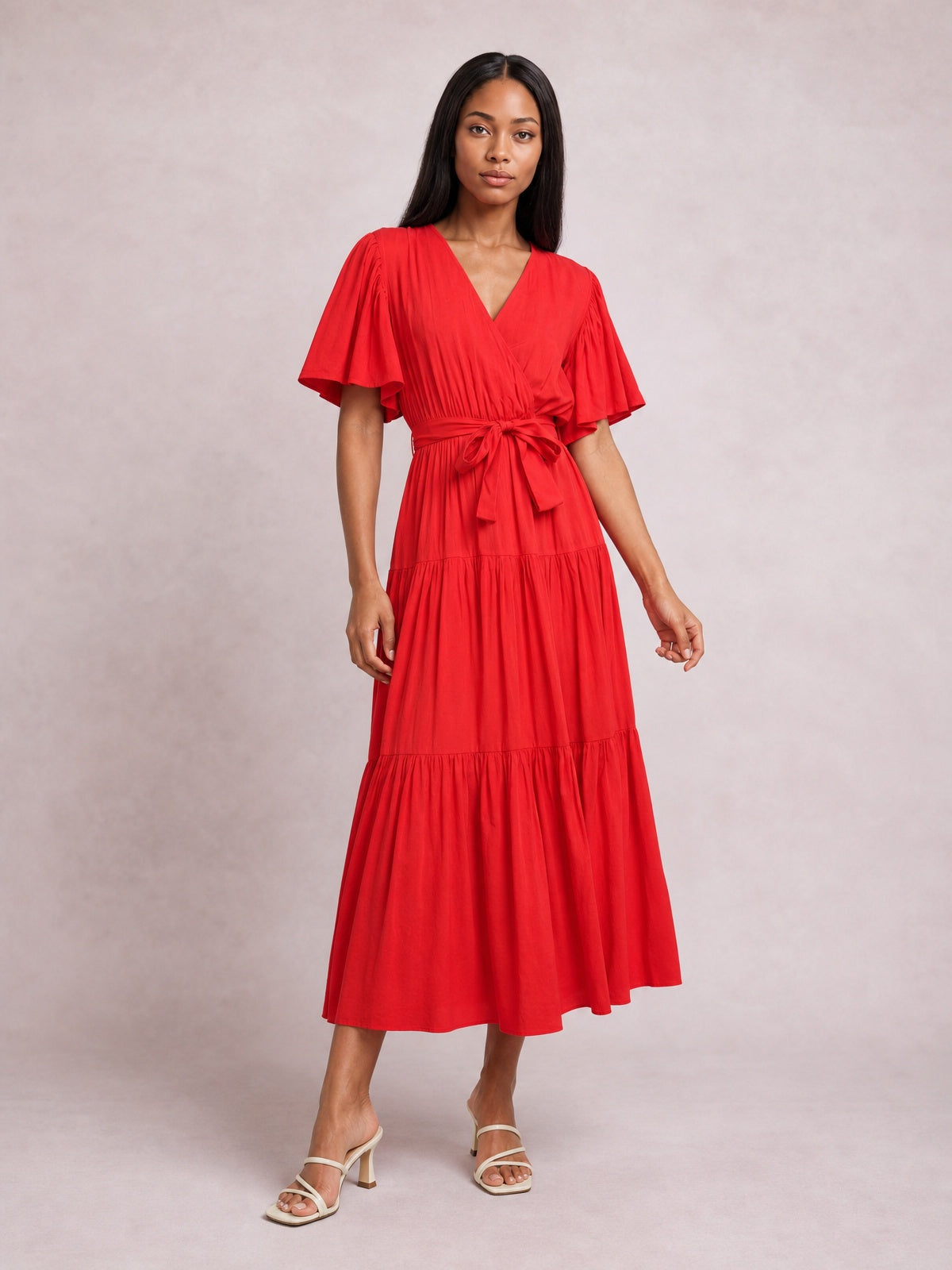 Model poses in a vibrant red front-tie midi dress with a V-neckline, flutter short sleeves, defined waist, and flowing tiered skirt.