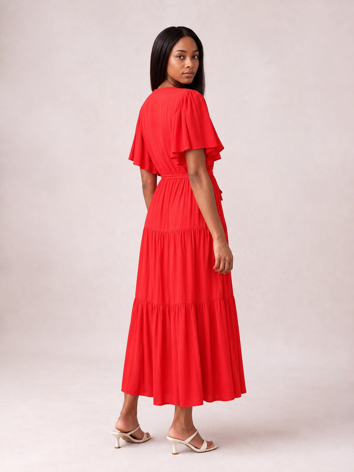 Model looks over her shoulder in a bright red tiered midi dress with flutter short sleeves, a gathered waist, flowing silhouette, and ankle-length hem.
