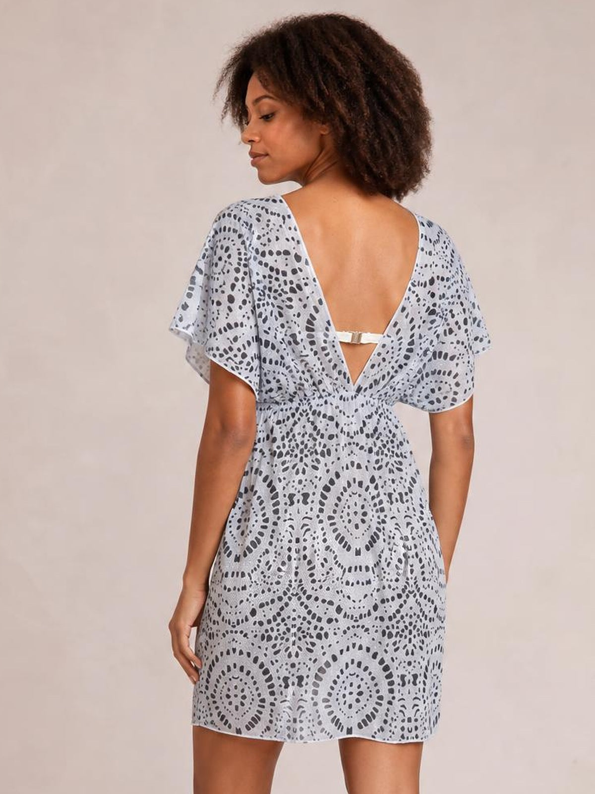 A model turns to show a light blue boho print beach cover-up dress with flutter sleeves, a loose short silhouette, and a deep V-back with strap detail.