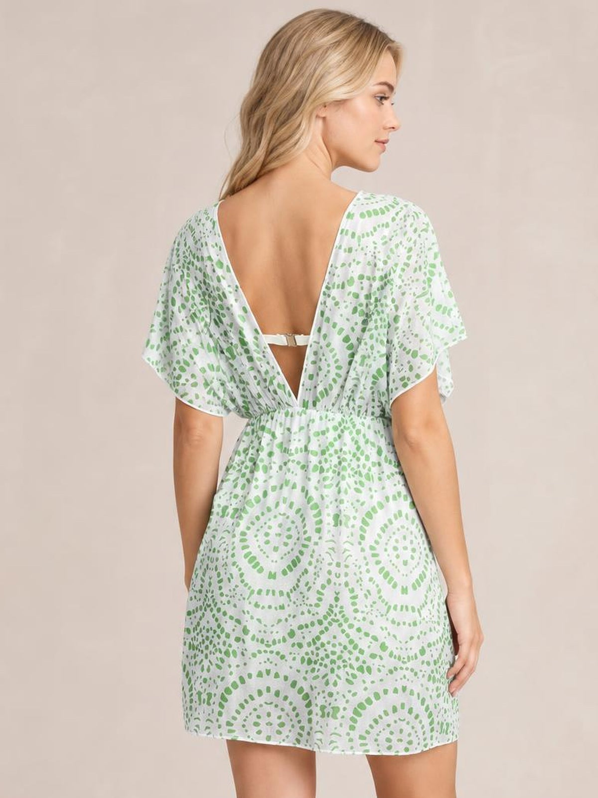 A model looks over her shoulder wearing a white and green boho print surplice beach cover-up dress with flutter sleeves, a deep back V, cinched waist, and short flowing silhouette.