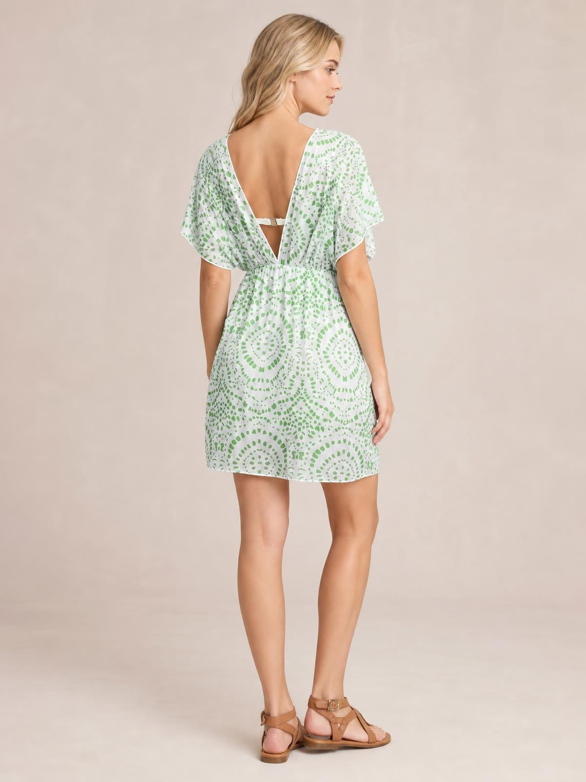 Model turns to show a white and green boho print beach cover-up dress with flutter sleeves, a low open back and strap, gathered waist, and loose short silhouette.