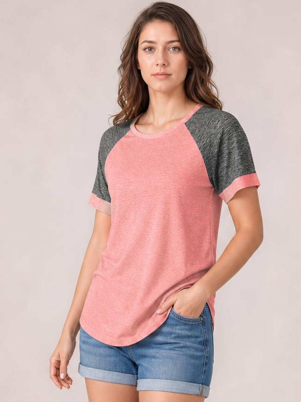 Model wearing a pink and charcoal contrast round neck short sleeve T-shirt with raglan-style construction, relaxed silhouette, and curved hem, hand in pocket.