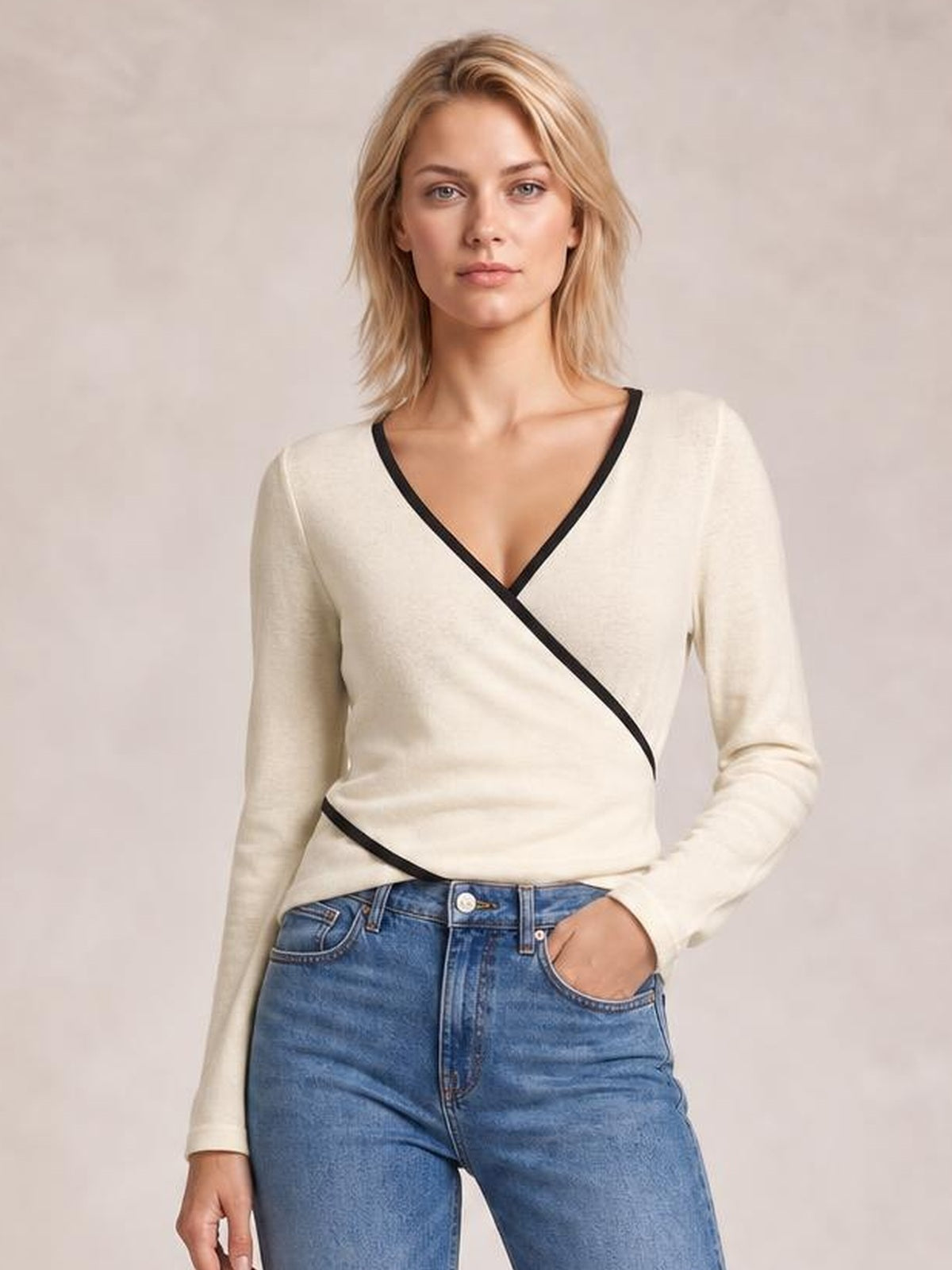 Model wearing an ivory long sleeve T-shirt with black contrast trim, V-neckline, and crisscross wrap-style detailing, posed with one hand in her jeans pocket.
