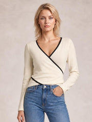 Model wearing an ivory long sleeve T-shirt with black contrast trim, V-neckline, and crisscross wrap-style detailing, posed with one hand in her jeans pocket.