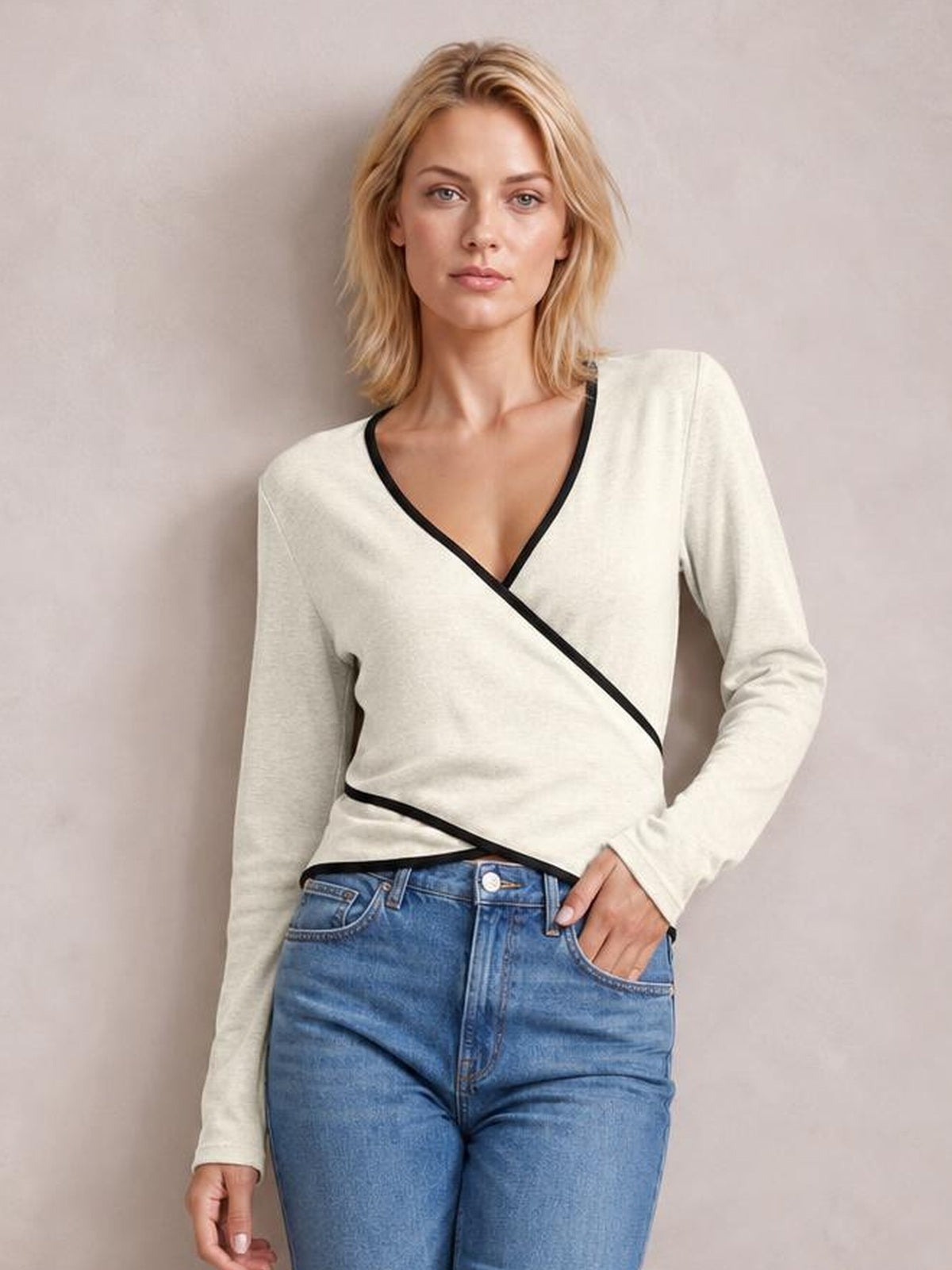 A model poses in a cream long-sleeve T-shirt with black contrast trim, a crisscross wrap front, V-neckline, and fitted silhouette, hand in her jeans pocket.