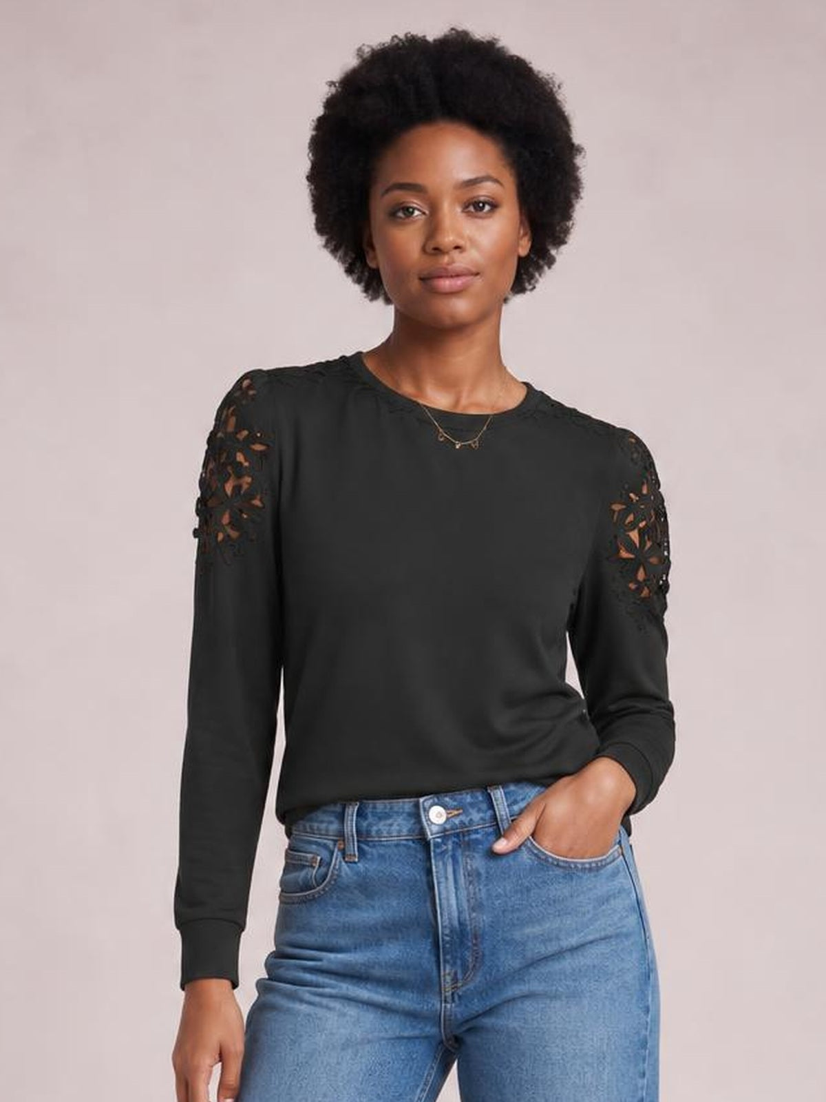 Model wearing a black cutout round neck long sleeve T-shirt with lace-detailed shoulders, a relaxed fit, and ribbed cuffs, posing with one hand in her jeans pocket.