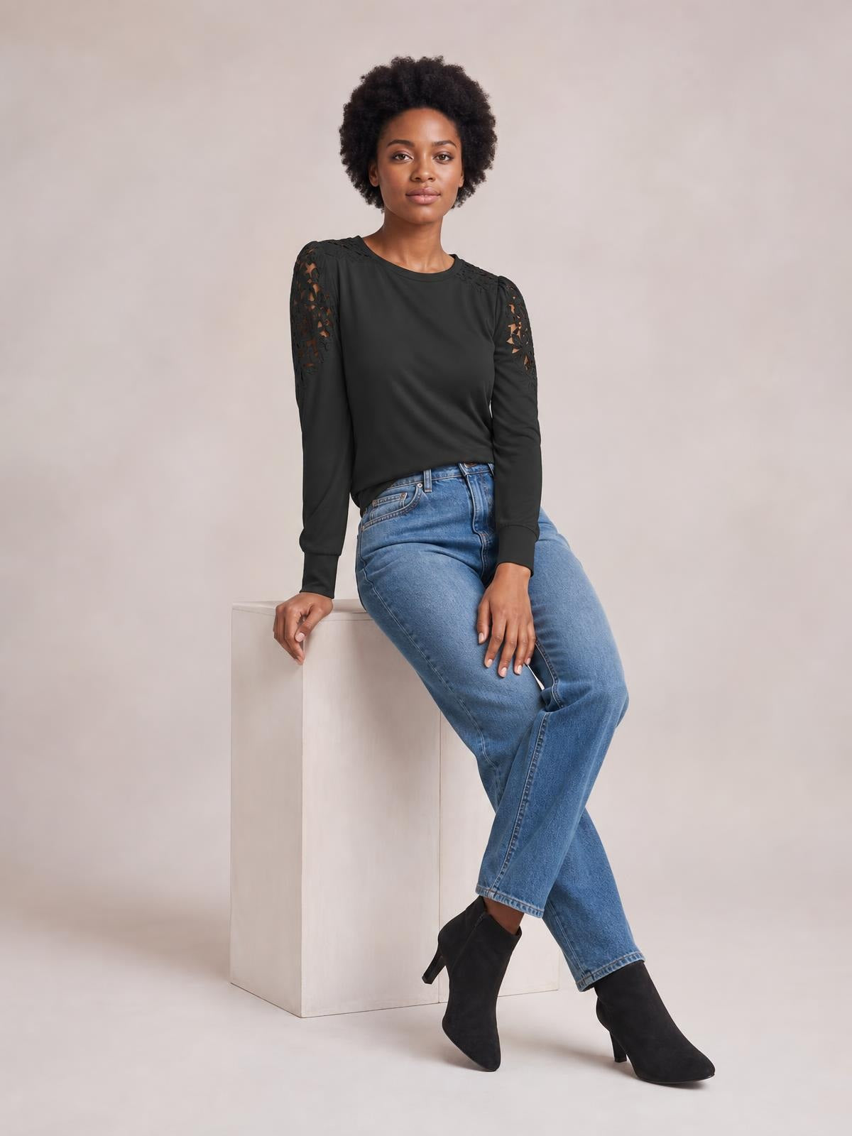 A seated model wears a black round-neck long-sleeve T-shirt with lace cutout panels on the shoulders and upper sleeves, creating a sleek silhouette.