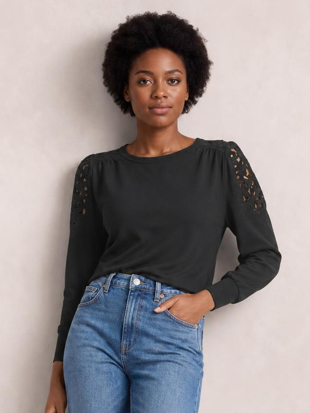 A model poses with one hand in her jeans pocket, wearing a black cutout round neck long sleeve T-shirt with sheer lace shoulder details and a relaxed fit.
