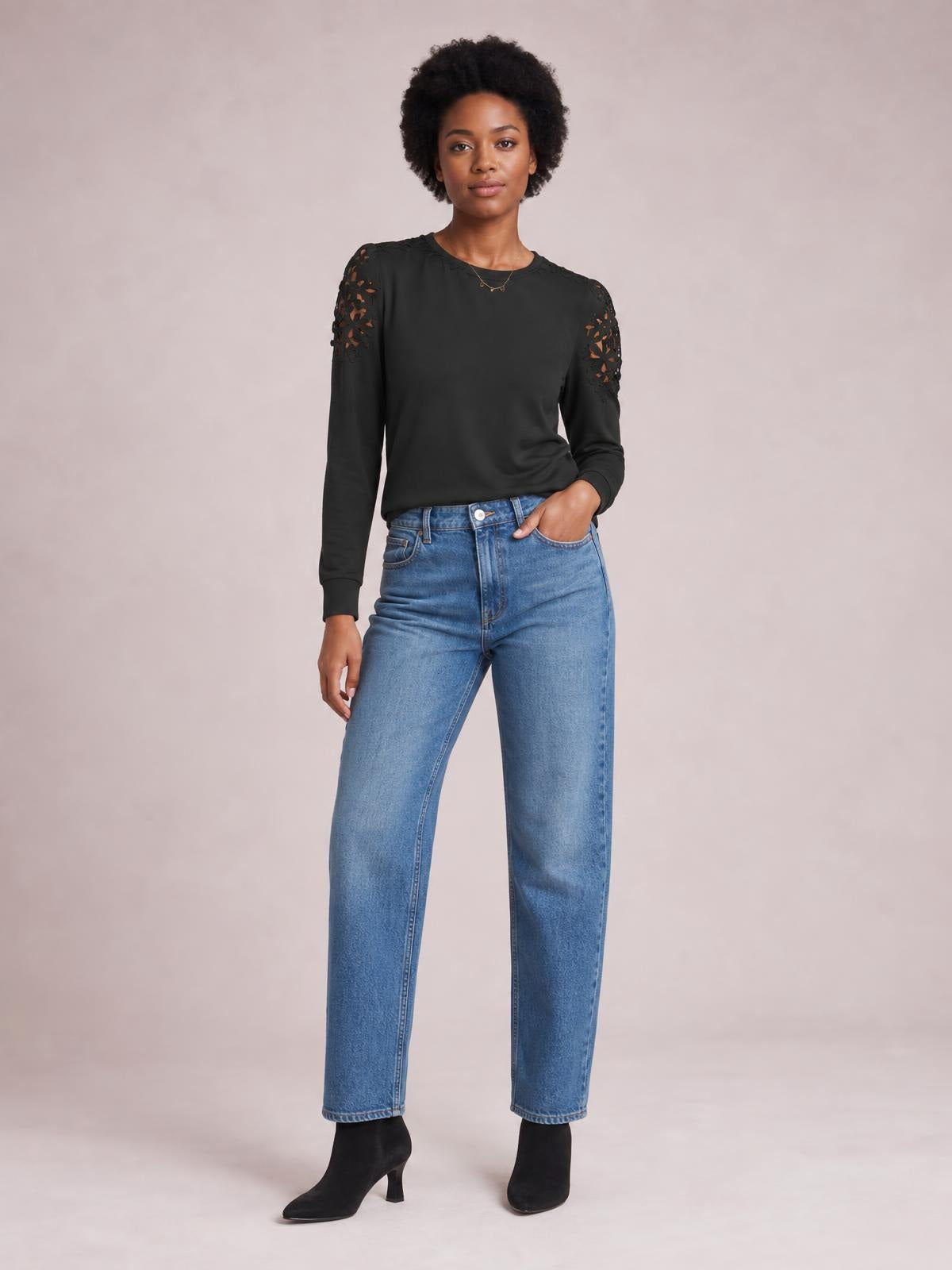Model wears a black cutout round neck long sleeve T-shirt with lace shoulder details and a relaxed silhouette, posing with one hand in her jeans pocket.