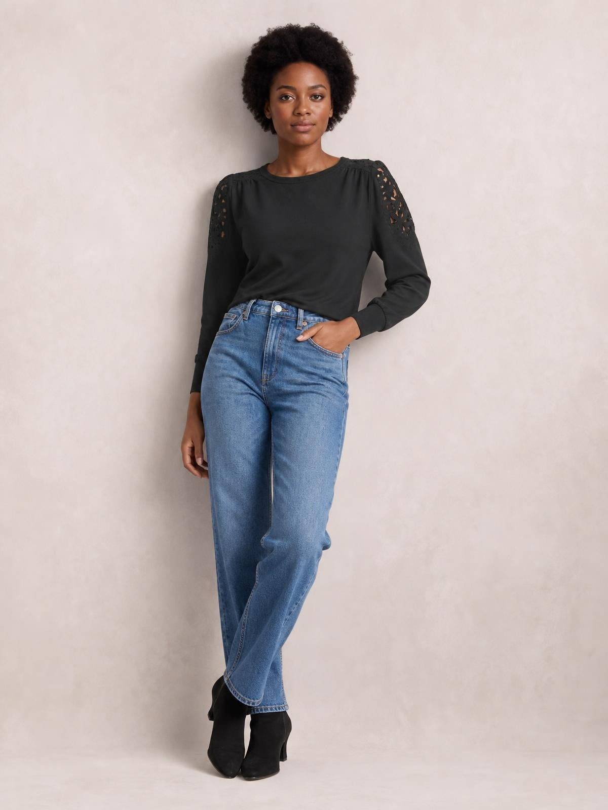 Model poses in a black cutout round neck long sleeve T-shirt with lace shoulder details and a relaxed silhouette, paired with jeans.