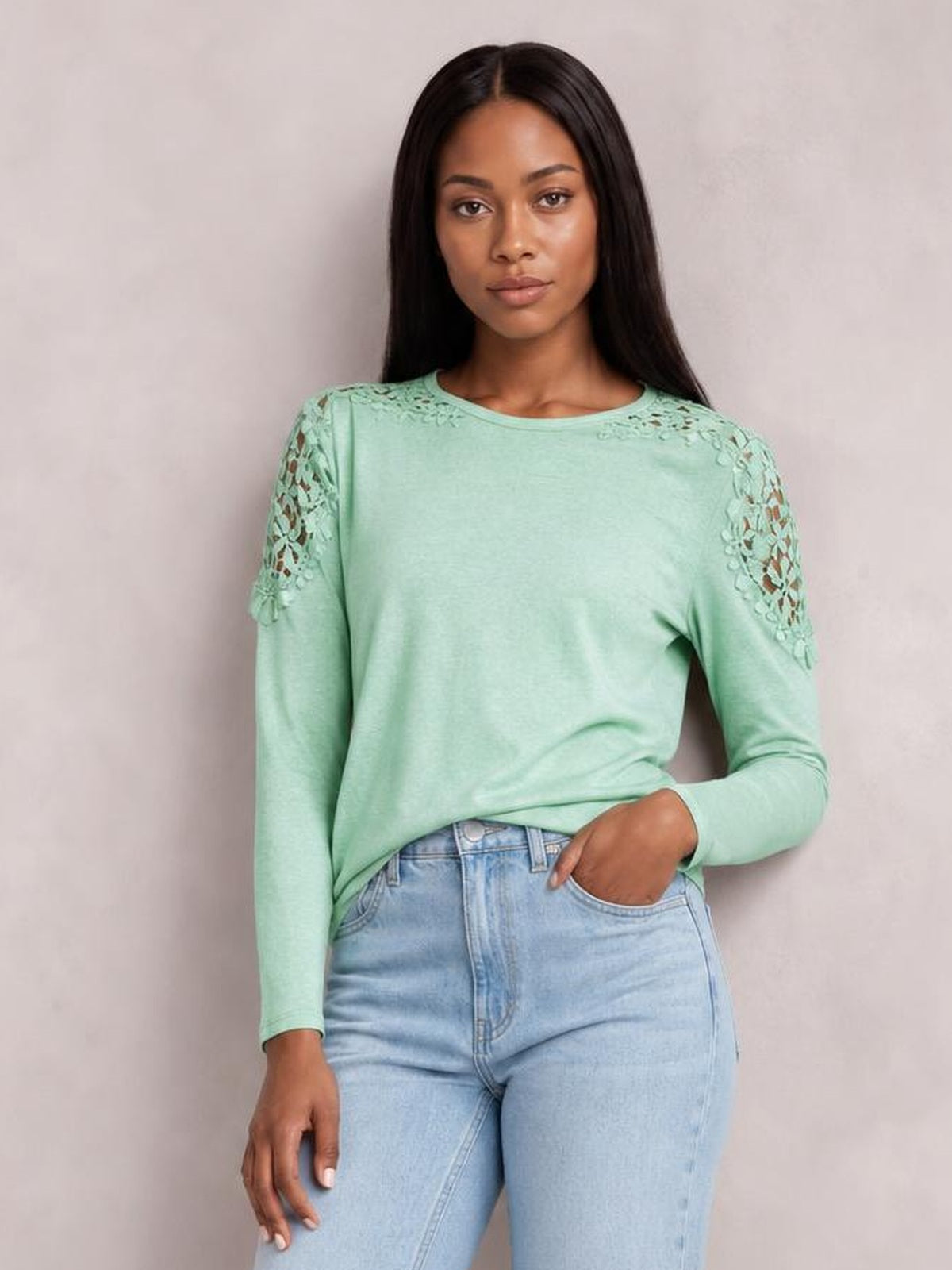 Model poses with one hand in her jeans pocket, wearing a mint green cutout round neck long sleeve T-shirt with floral lace shoulder details and a relaxed silhouette.