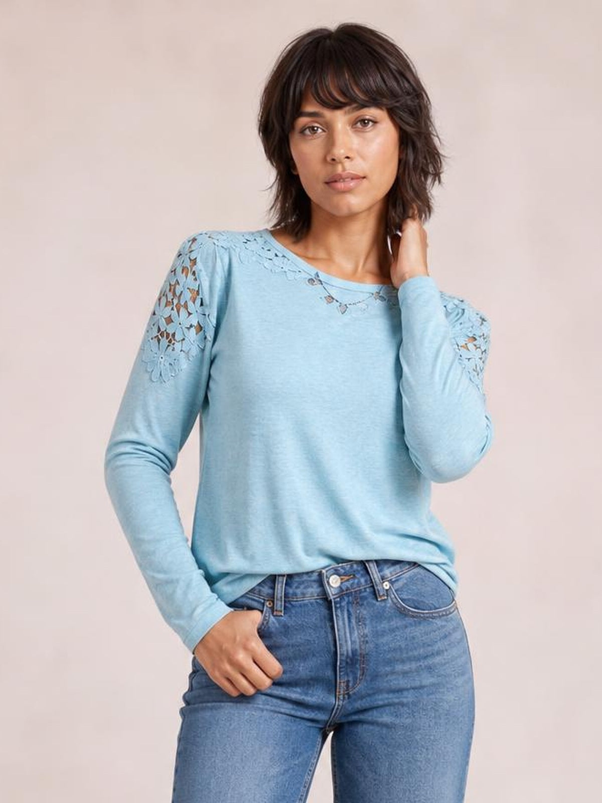 A model poses in a light blue cutout round-neck long-sleeve T-shirt with lace-detail shoulders and sleeves, a relaxed silhouette, and one hand in her pocket.