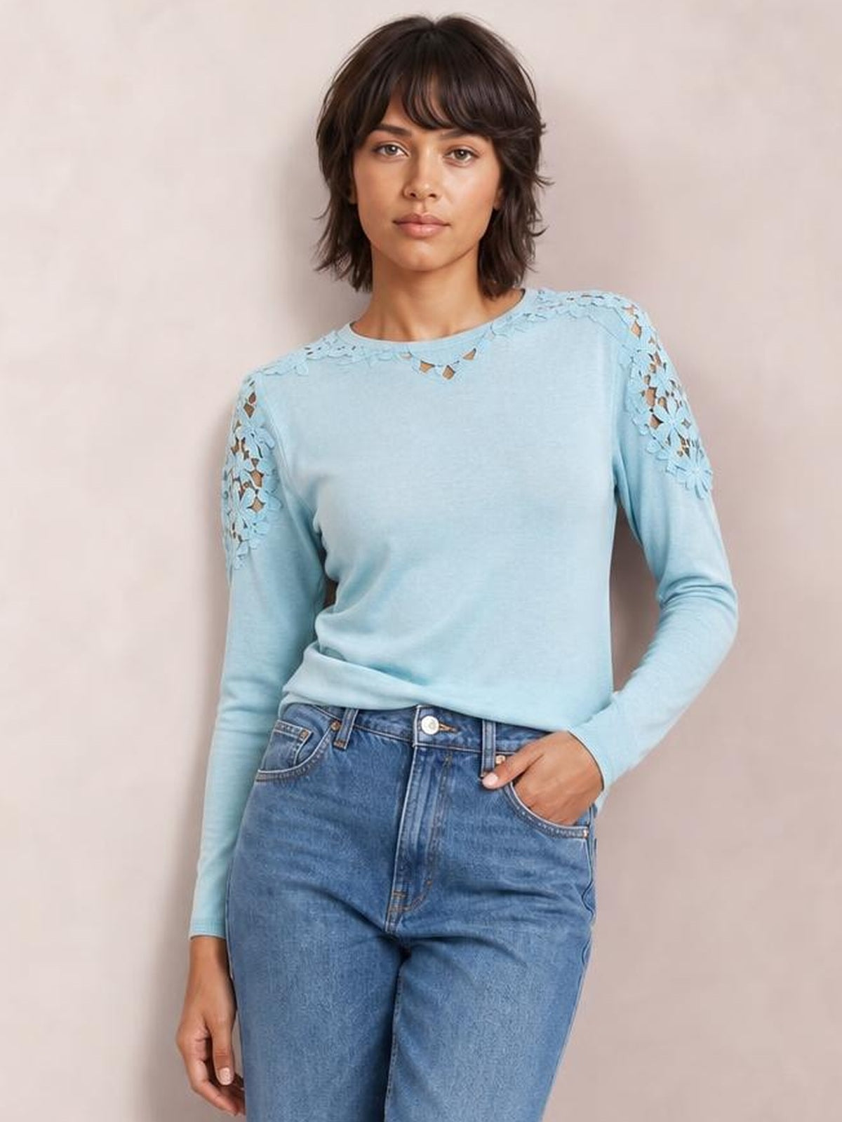 A model wears a light blue round-neck long-sleeve T-shirt with lace cutout detailing at the shoulders and neckline, styled in a relaxed fit with one hand in her jeans pocket.