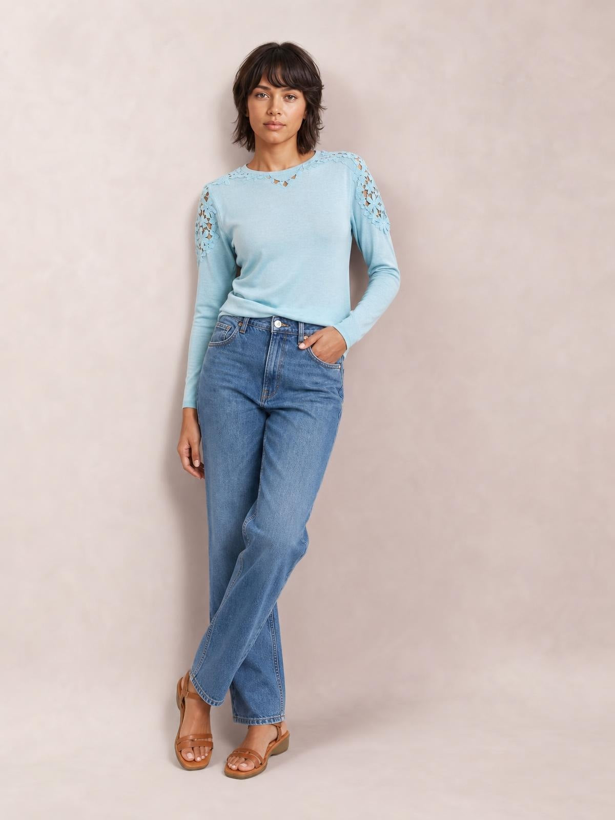 A model poses with one hand in her jeans pocket wearing a light blue round-neck long-sleeve T-shirt with lace cutout shoulder details and a softly fitted silhouette.