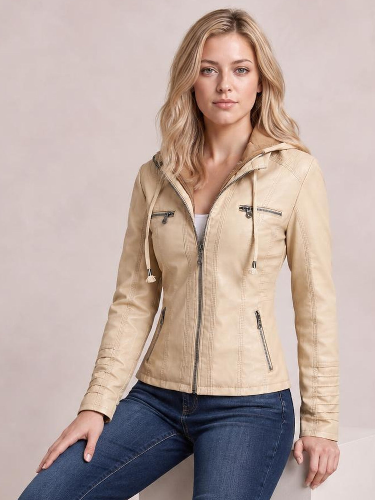 A seated model wears a beige faux leather moto jacket with a hood, full zip closure, seam detailing, chest and side zip pockets, and drawstrings.