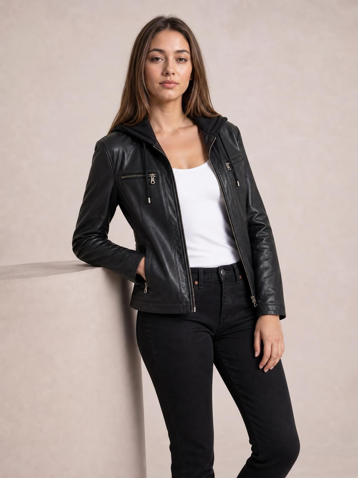Model poses with one hand in the pocket wearing a black faux leather moto jacket with hood, zip closure, seam detailing, and multiple zippered pockets.