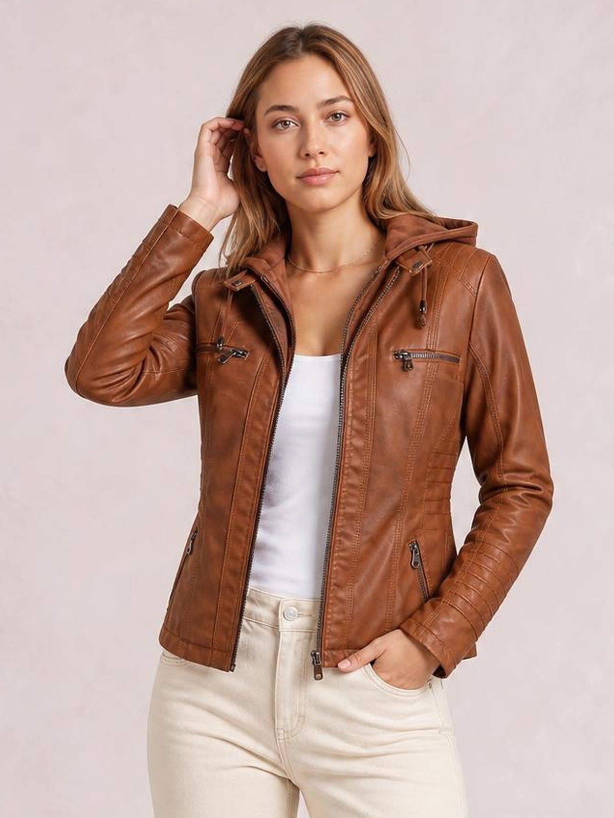 Model poses in a brown faux leather moto jacket with a hood, zip closure, seam detailing, and multiple zippered pockets in a regular fit.