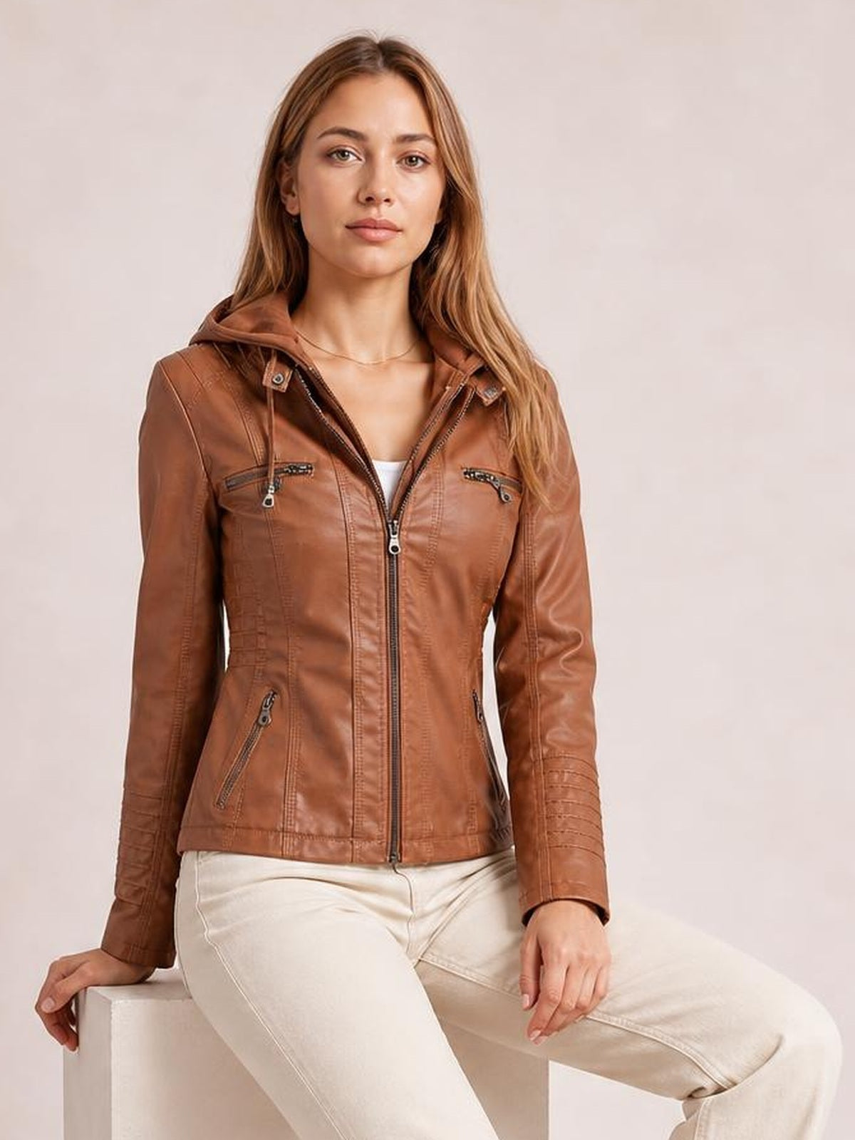 Model sits with one hand resting beside her, wearing a brown faux leather moto jacket with hood, zip closure, seam detailing, and multiple zippered pockets.