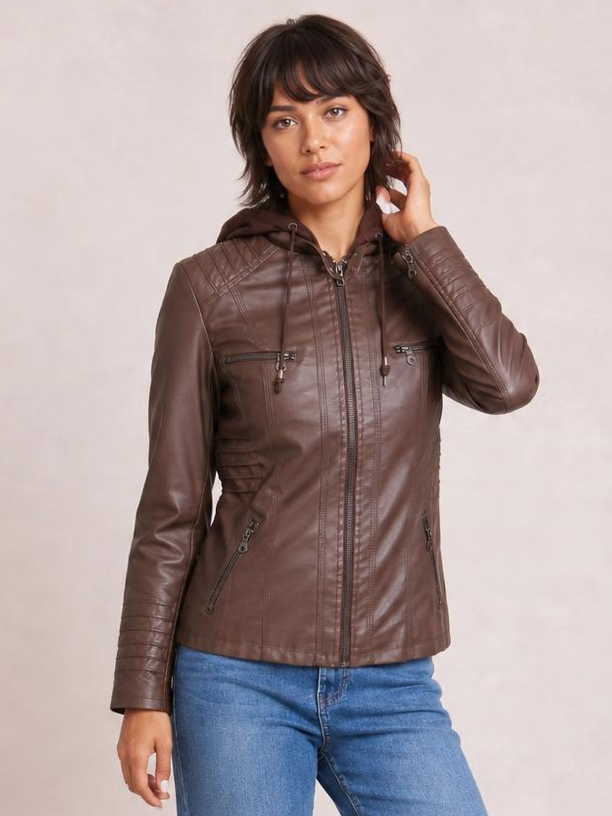Model poses in a brown faux leather moto jacket with a zip-up front, drawstring hood, seam detailing, and multiple zippered pockets.