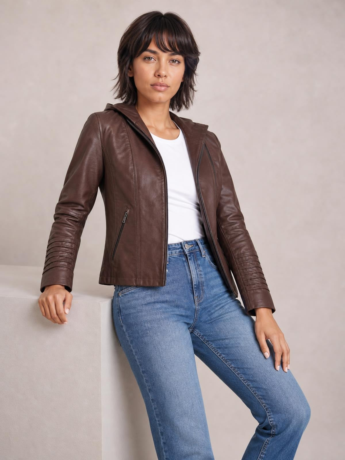 Model leans against a ledge wearing a brown faux leather moto jacket with hood, zip closure, seam detailing, zip pockets, and a regular silhouette.