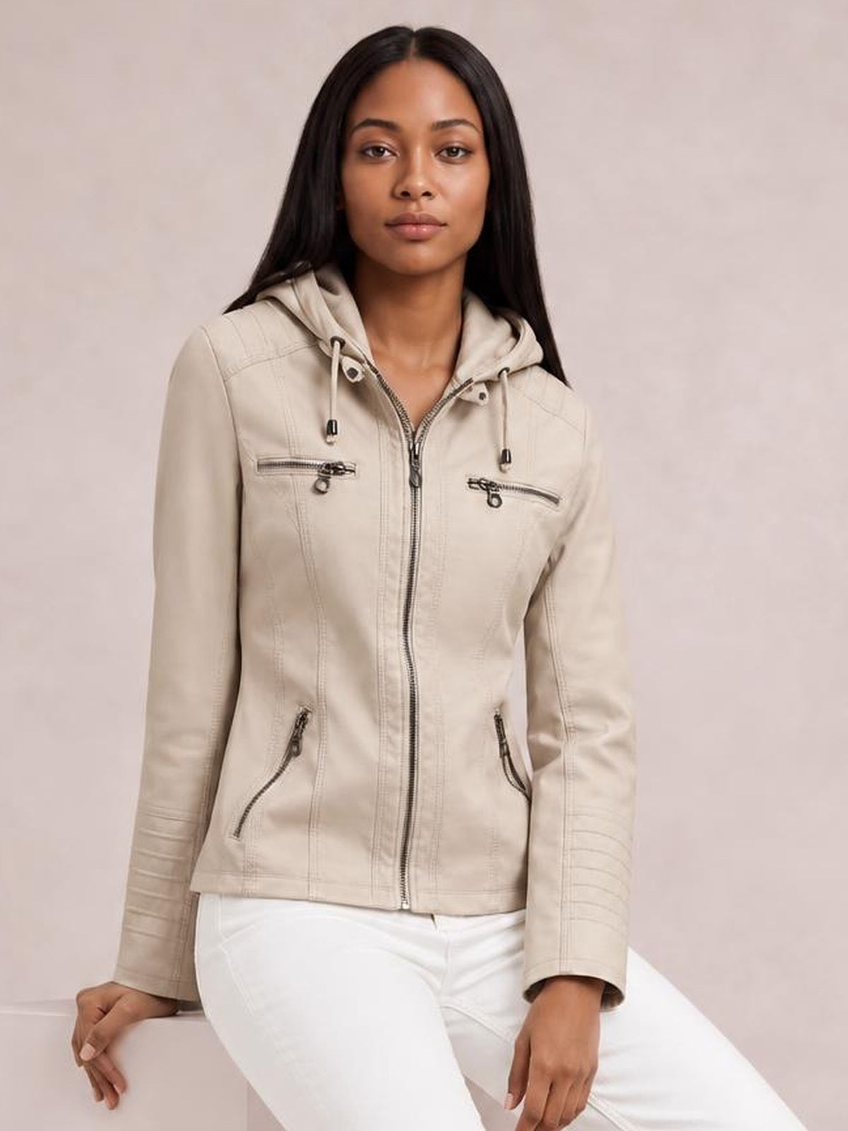 Model seated in a light beige faux leather moto jacket with a drawstring hood, zip closure, seam details, and multiple zippered pockets.